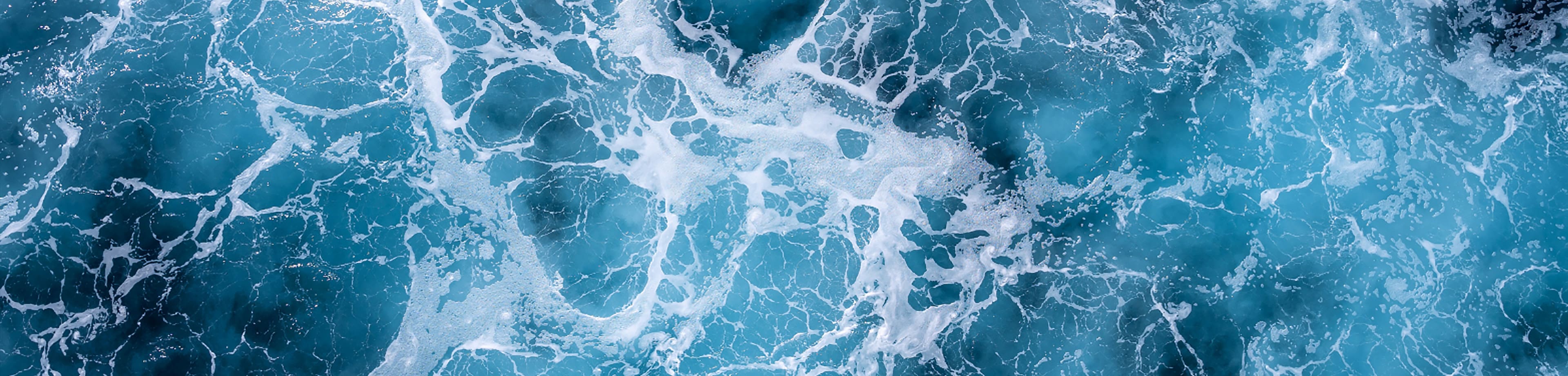 An aerial view of ocean water in motion, showcasing swirling waves and foam patterns on the surface.